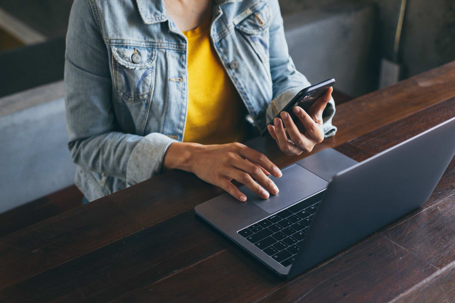 A woman scrolling on a laptop with one hand while holding her cell phone in the other hand.